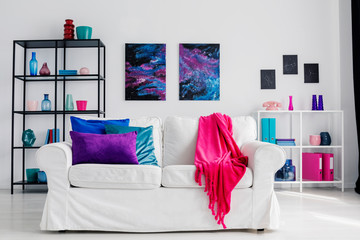 Vertical view of stylish living room with comfortable white couch with pink blanket and blue and purple pillows, cosmos graphics on the wall and metal shelves with accessories, real photo