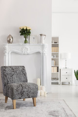 Flowers on fireplace portal in white apartment interior with grey patterned armchair on carpet....