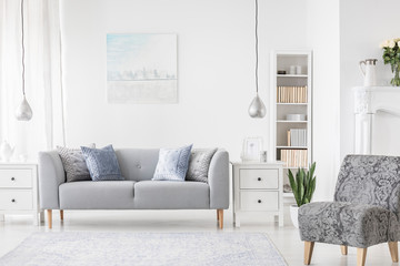Lamp above white cabinet between plant and grey sofa in simple flat interior with armchair. Real photo