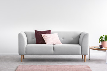 Purple and pastel pink pillow on the grey comfortable couch in bright living room interior with pink carpet and coffee table, real photo with copy space on the empty white wall