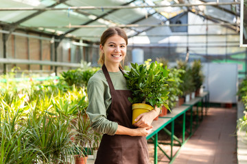 Image of caucasian woman gardener 20s wearing apron standing with plants in hands, while working in greenhouse