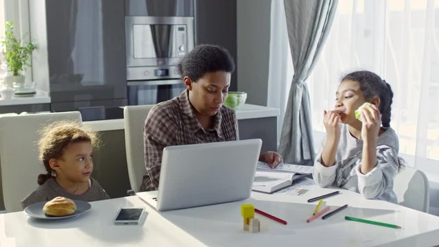 PAN of black woman with laptop sitting at kitchen table and writing notes in planner as her daughters having snacks: preschool age girl eating bun and looking at elder sister chewing green apple