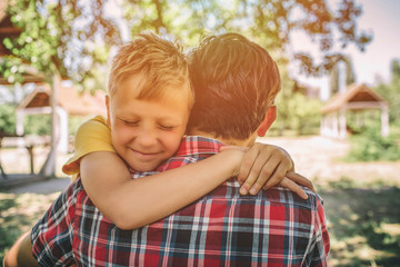 Fototapeta premium Cheerful and happy boy is hugging his father. Boy is keeping eyes closed and smiling. They are standing outside in park.