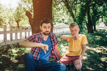 Sad and upset guys are sitting on knees and holding their big thumbs down. Boys are looking on camera with serious sight. They are sitting in park.