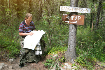Tourist with a map near the signs. Inscriptions Tungur village, Kara Turek mountain. A man traveler got lost in a hike and is looking for a way on the map. - obrazy, fototapety, plakaty