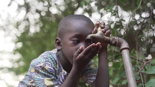 Candid shot of an African black baby boy outdoors drinking clean fresh safe water from a tap. 4K RAW clip, please modify and edit (color grade, stabilize, etc.) in postproduction.
