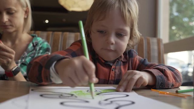 Child draws with crayons on a piece of paper in cafe. Preschool education