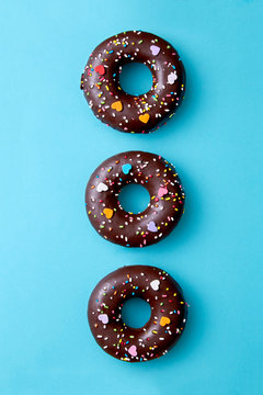 Three chocolate donuts alligned on a blue background viewed from above. Sweet donuts sprinkled with colorful decoration. Top view