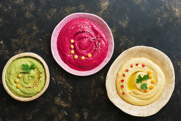 A variety of colored hummus, classic hummus, beet hummus, hummus with avocado on a dark rustic background. Top view, flat lay. Clean eating, dieting, vegetarian party food