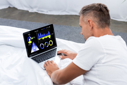 rear view of adult man laying in bed and using laptop with infographic on screen at home