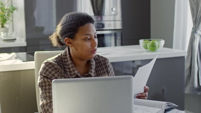 PAN of focused black woman working from home: she sitting at kitchen table and looking at document, then typing on laptop and writing notes in planner