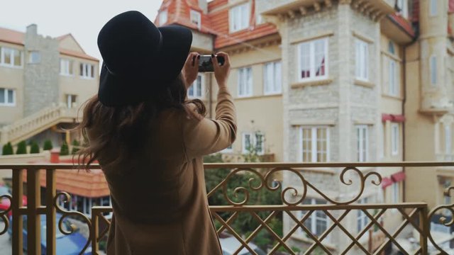 View from the back, close-up, steadikam slender woman in a coat and hat walks around the city, takes a photo of the architect on her smartphone, enjoys a walk. The girl flew over the weekend to Europe