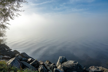Soft ripples in misty landscape. Peaceful thick fog on lake. Rocks and tree in foreground. Sweden