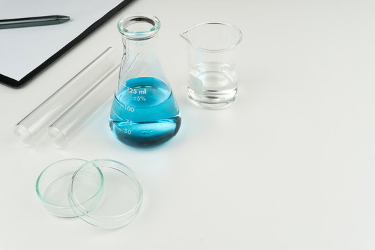 Laboratory Equipments. A Blue Liquid Flask, Clear Liquid Beaker, Test Tubes, Petri Dish And A Clipboard With Pen On The White Table