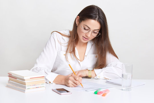 Female entrepreneur writes organisation plan, sits at desktop, uses books and pencil, drinks fresh water from glass, concentrared on work, wears round optical glasses, isolated over white background
