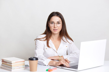 Succesful copywriter busy working with laptop computer, wears round glasses, drinks takeaway coffee, poses against white background, dressed in formal clothes. People, work and occupation concept