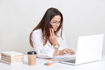 Skilled female student writes homework, works on laptop computer, uses application, keyboards and searches internet, has attentive look into screen, stay at table indoor, books and coffee near