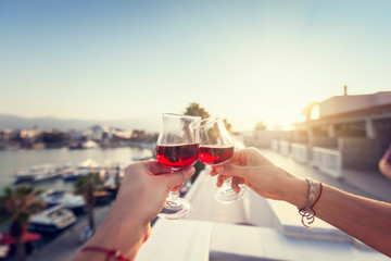 Two glasses with red wine in human hands against the background of the resort city and sunset,...