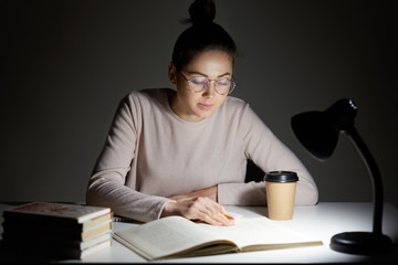 Busy female teenager reads book, uses table lamp, prepares for final examination, sits at desktop,...