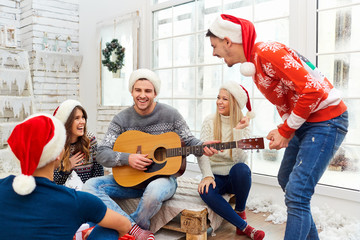 A group of friends in Santa's hats are playing the guitar and singing at home on Christmas Day.