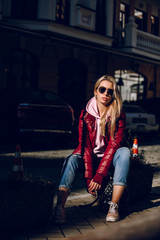 Obraz na płótnie Canvas Street fashion concept - portrait of a beautiful girl sitting outside, blue jeans, sunglasses, hard lights, red jacket, autumn weather