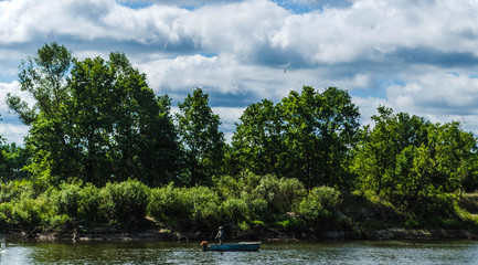 View of the river where a fisherman in a boat catches fish on a background of green trees and blue sky. Belarusian Polesie. Pripyat National Park