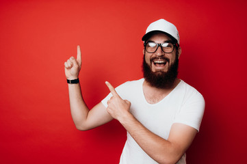Cheerful bearded hipster man wearing eyeglasses and pointing at copyspace over red background