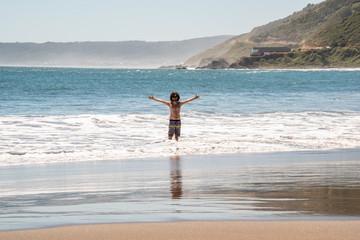 Young man with open arms with his legs in the cold Pacific Ocean and a mountain range on the horizon