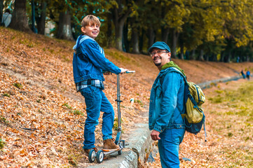 Happy dad with son in jeans clothes are walking along the autumnal embankment with a scooter