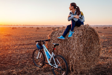 Young bicyclist having rest after a ride in autumn field at sunset. Woman admiring view sitting on...