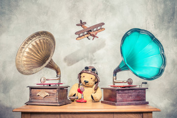 Teddy Bear with aviator's helmet and goggles, aged antique gramophone phonograph turntables with old retro microphone, flying toy plane with its shadow on concrete wall. Vintage style filtered photo