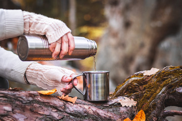 Woman with knitted glove is pouring a hot drink into mug from thermos. Refreshment during hiking. 