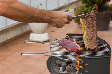 Man turning steak around on barbecue grill with big fork. Charcoal and fire flames to cook raw...