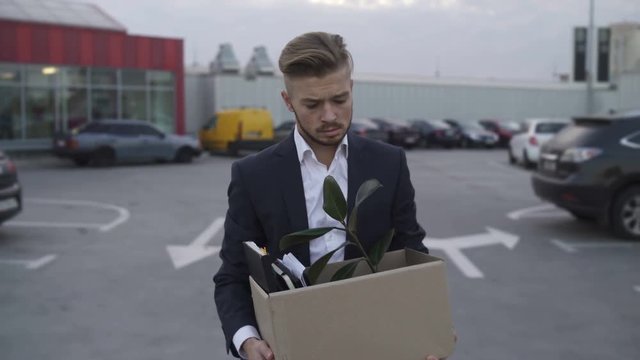 Emotional disappointed Corporate worker gets fired and walking down parking lot at the Roof with carton box and documents with a table flower. Lost His Job beacause a mistake.