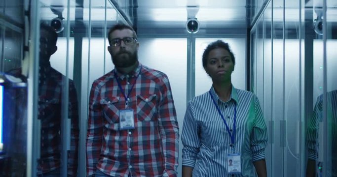 View of adult black woman and bearded man walking in corridor among server racks in data center