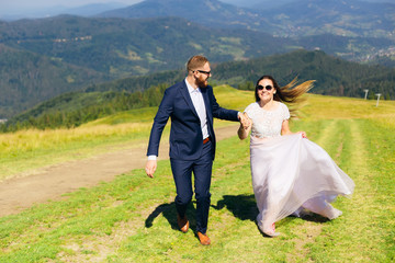 the groom in a stylish suit and a beard and sunglasses holds the bride by hand and they run on the grass on top of the mountain