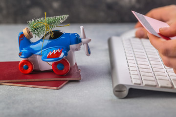 Small airplane with christmas tree, passports, keybaord, credit card on grey background. Vacation, tourism, travel for christmas. Woman hands booking tickets online for Christmas eve.