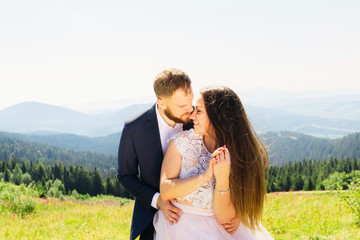 the groom embraces the bride behind the waist from the back and the bride in white dress with lace closed her eyes on the background of the mountains