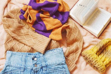 Flat lay autumn and winter fashion photo. Brown woolen sweater, striped scarf, blue denim skirt, woolen hat and book on a bed