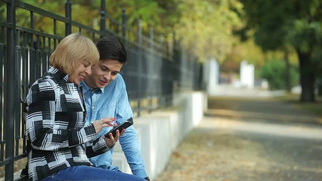  Optimistic view of a cheery blond woman giving high five to her sportive brunet son sitting on a fence parapet and surfing the net in autumn  
