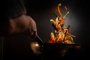 Colorful mix of vegetables being cook in a wok pan in a dark environment,  while the chef is throwing them up. - obrazy, fototapety, plakaty