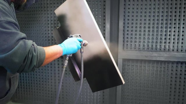 Man applying black anti-corrosion powder coating to metal sheet using a spray gun in factory.