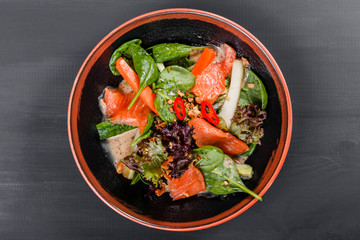 Leaf vegetable salad with smoked salmon, spinach, peppers, cucumbers and sauce on plate on dark wooden background.
