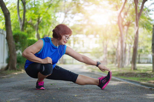 Senior asian woman stretch muscles at park and listening to music. Athletic senior exercising together outdoor. Fit senior runners stretching before running outdoors.