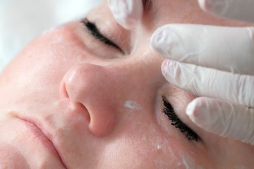 Close-up. The hands of a beautician or a doctor in white gloves put eye cream on the face of a full woman. Skin cleaning in the cosmetology center. Cosmetological therapeutic beauty procedure.
