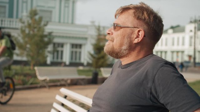 Portrait of a mature man with beard and glasses relaxing outdoors sitting on a park bench. 