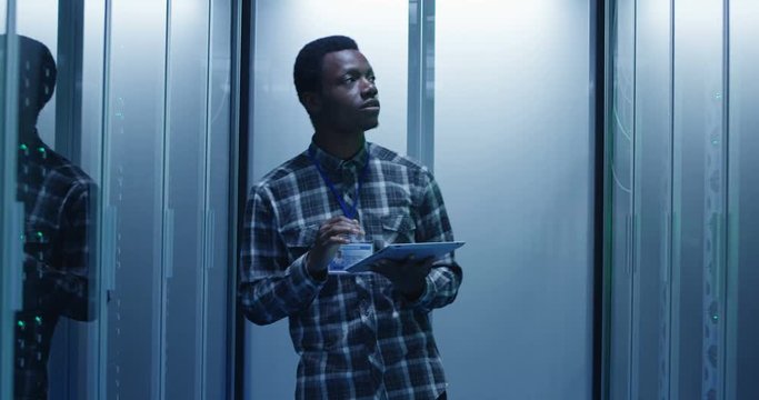 Young African American IT engineer using tablet and walking in corridor with server rack setting hardware in data center and check server hardware