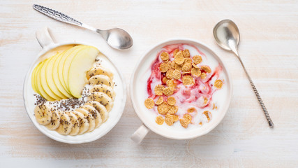 Two greek yogurt with jam, apple, muesli, chia seeds and banana in white bowl on white wooden table, top view, banner