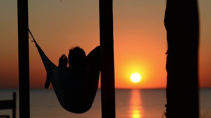 Man relaxes in a hammock on a bungalow veranda, admiring the sunset over the sea in front of him. Amazing sunset on the beach. Back view. - Powered by Adobe