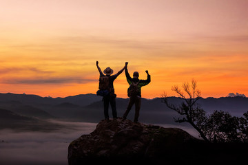 Fototapeta premium Two tourists with backpacks in the campaign raise their hands up on morning foggy mountain peak. Panoramic view of tourist on mountains hill raising his hands. Successfully achieving your goal concept